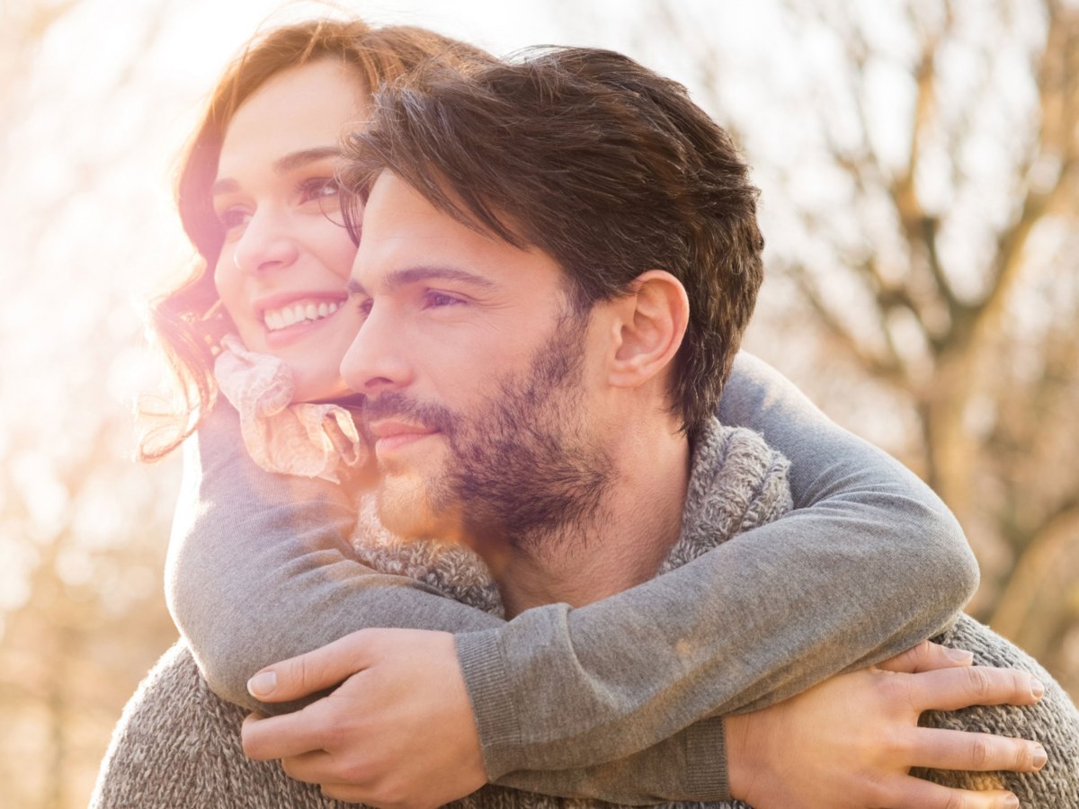 Frau umarmt Mann von hinten, beide schauen nach links und lächeln.