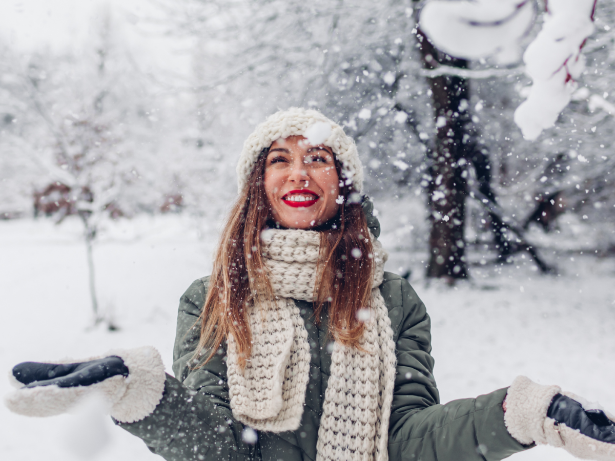 Junge Frau mit roten Lippen, Wollm&uuml;tze und Schal lacht strahlend in fallendem Schnee vor winterlicher Parklandschaft.