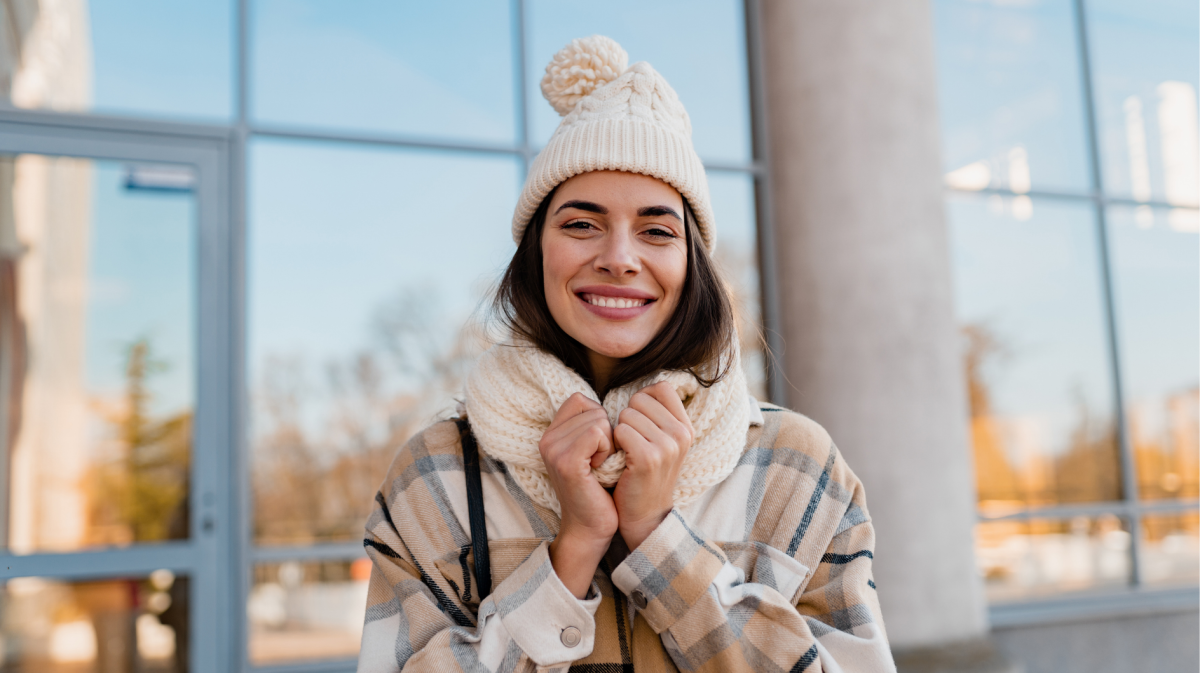 Fröhliche Frau mit beigem Winteroutfit hält Schal fest, sonniger Tag, reflektierende Fenster im Hintergrund.