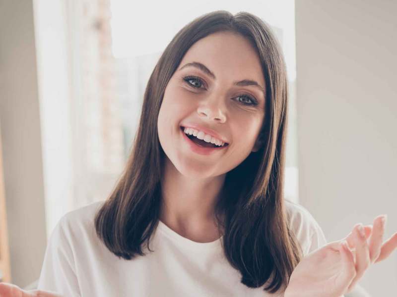 Eine junge Frau mit schulterlangem braunem Haar und weißem T-Shirt lächelt freundlich in die Kamera, sitzend in einem hellen Raum.