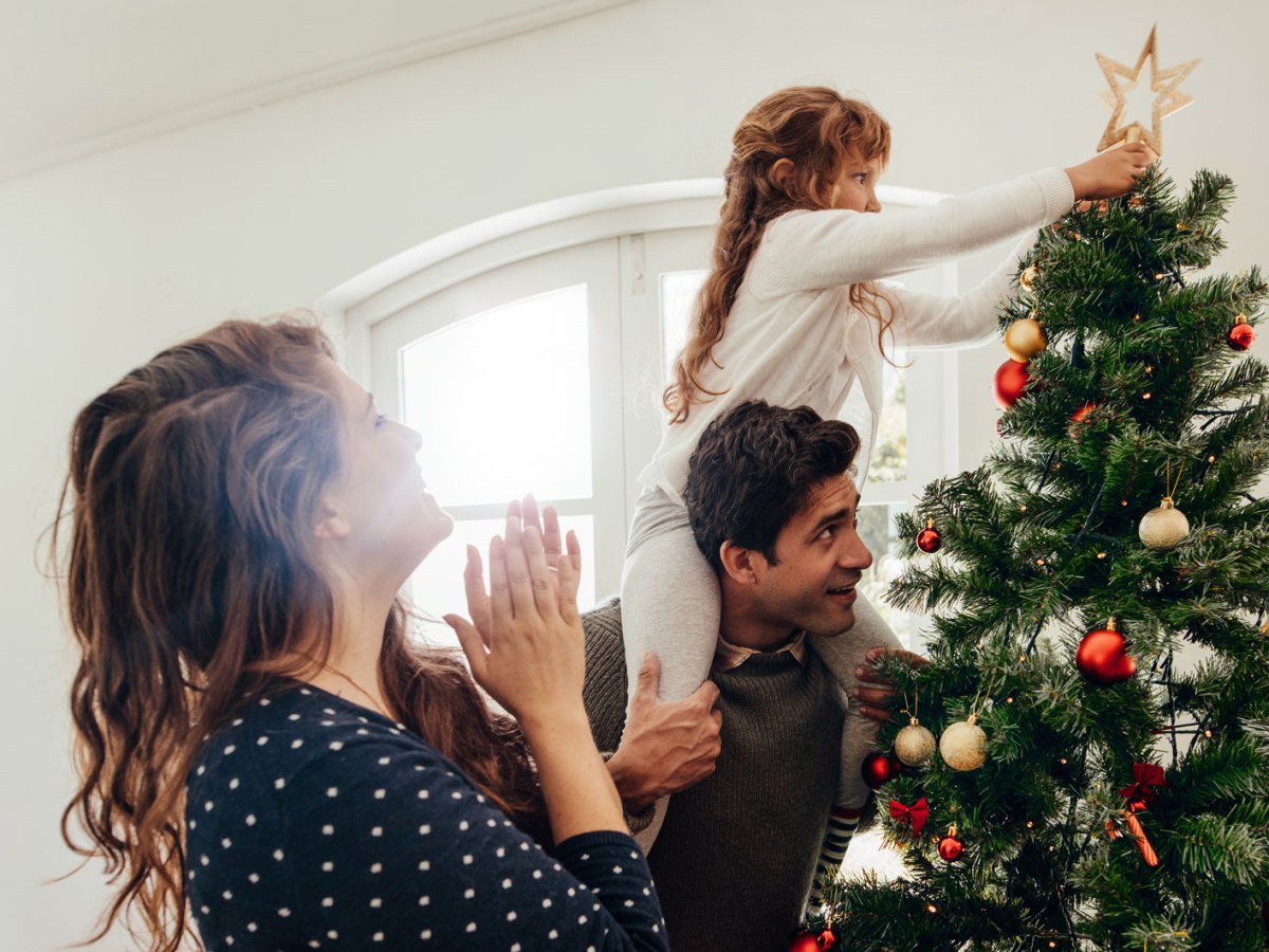 Mutter, Vater und Tochter haben den Weihnachtsbaum geschm&uuml;ckt. Die Tochter, auf den Schultern des Vaters sitzend, steckt dem Baum die Stern-Spitze an.