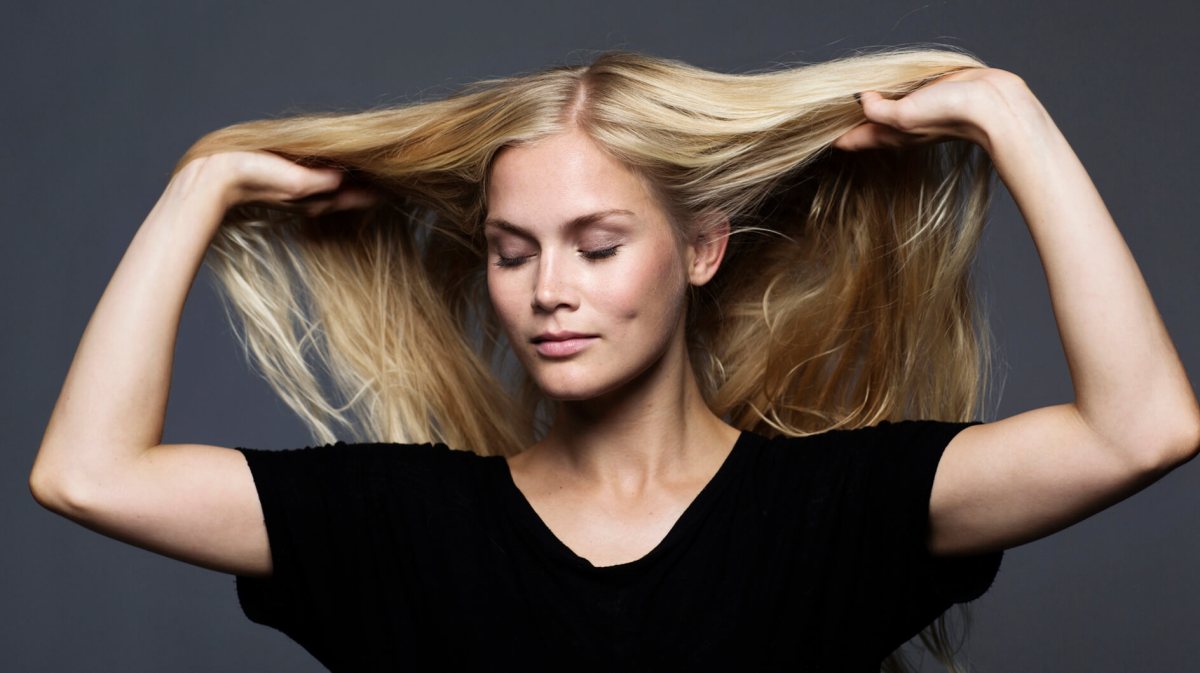 Junge blonde Frau in schwarzem Shirt hält ihr langes Haar mit geschlossenen Augen seitlich hoch vor dunkelgrauem Hintergrund.