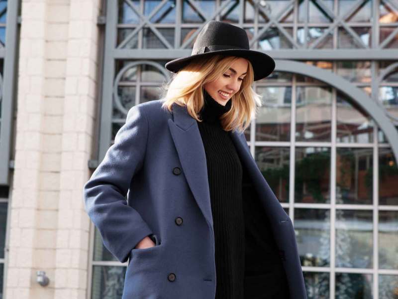 Junge Frau mit blonden Haaren und einem Hut und einem Mantel läuft durch die Stadt.