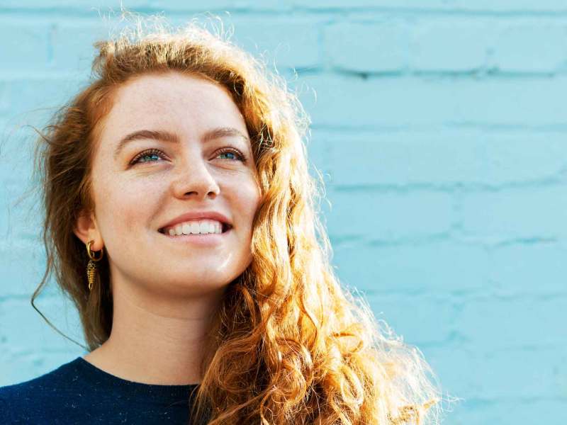 Frau mit roten, lockigen Haaren steht lächelnd vor einer hellblauen Wand.