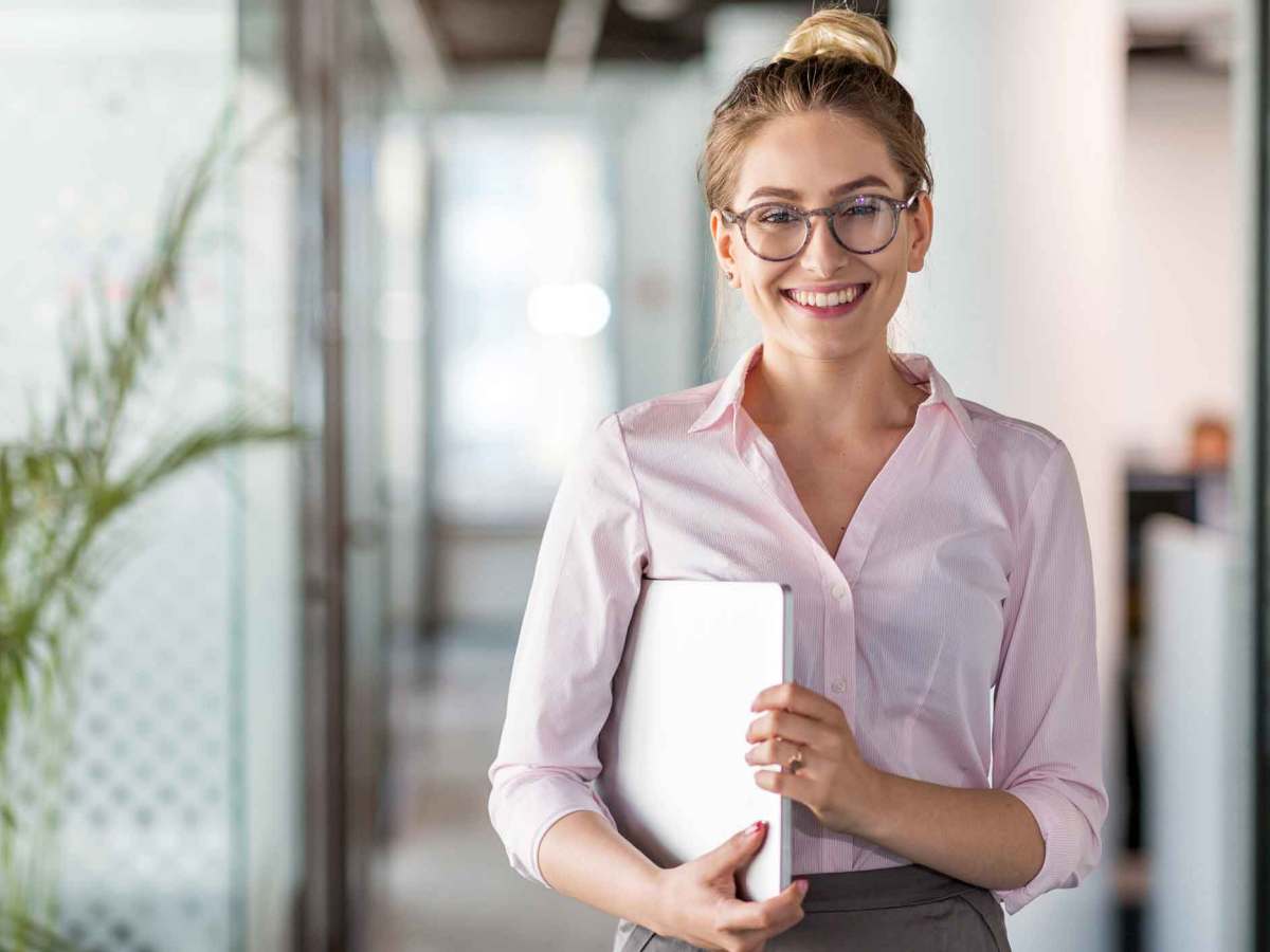 Frau mit Brille steht im B&uuml;ro und h&auml;lt ein Notebook unter dem Arm.