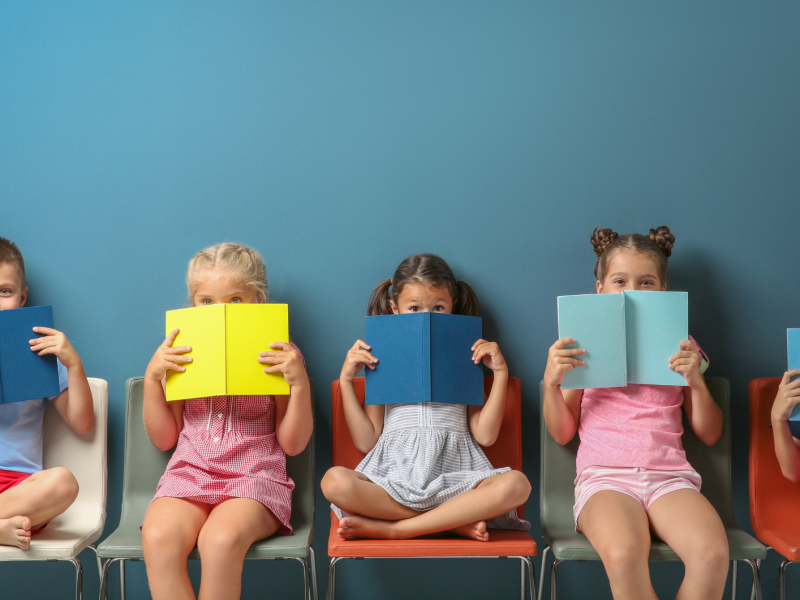 Kinder mit B&uuml;chern sitzen vor einer blauen Wand auf St&uuml;hlen