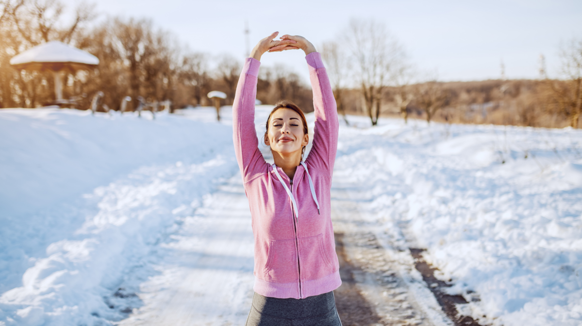 Eine junge Frau mit braunem Haar und rosa Kapuzenjacke streckt sich lächelnd auf einem verschneiten Weg in der Wintersonne.