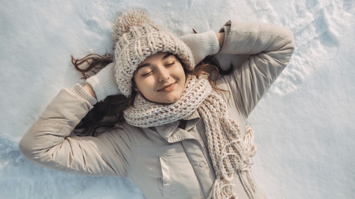 Glückliche Frau in beiger Winterjacke, Strickmütze und Schal liegt entspannt mit verschränkten Armen auf schneeweißem Boden.