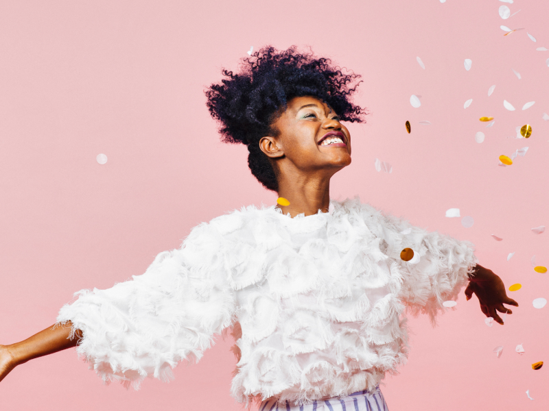 Junge Frau mit lockigem Afro und weißem Fransenshirt lacht strahlend mit ausgestreckten Armen, umgeben von Konfetti vor rosa Hintergrund.