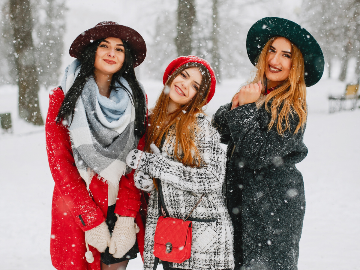 Drei Frauen im Schnee, mit Wintermantel und H&uuml;ten, die allesamt in die Kamera l&auml;cheln, w&auml;hrend es um sie herum schneit.