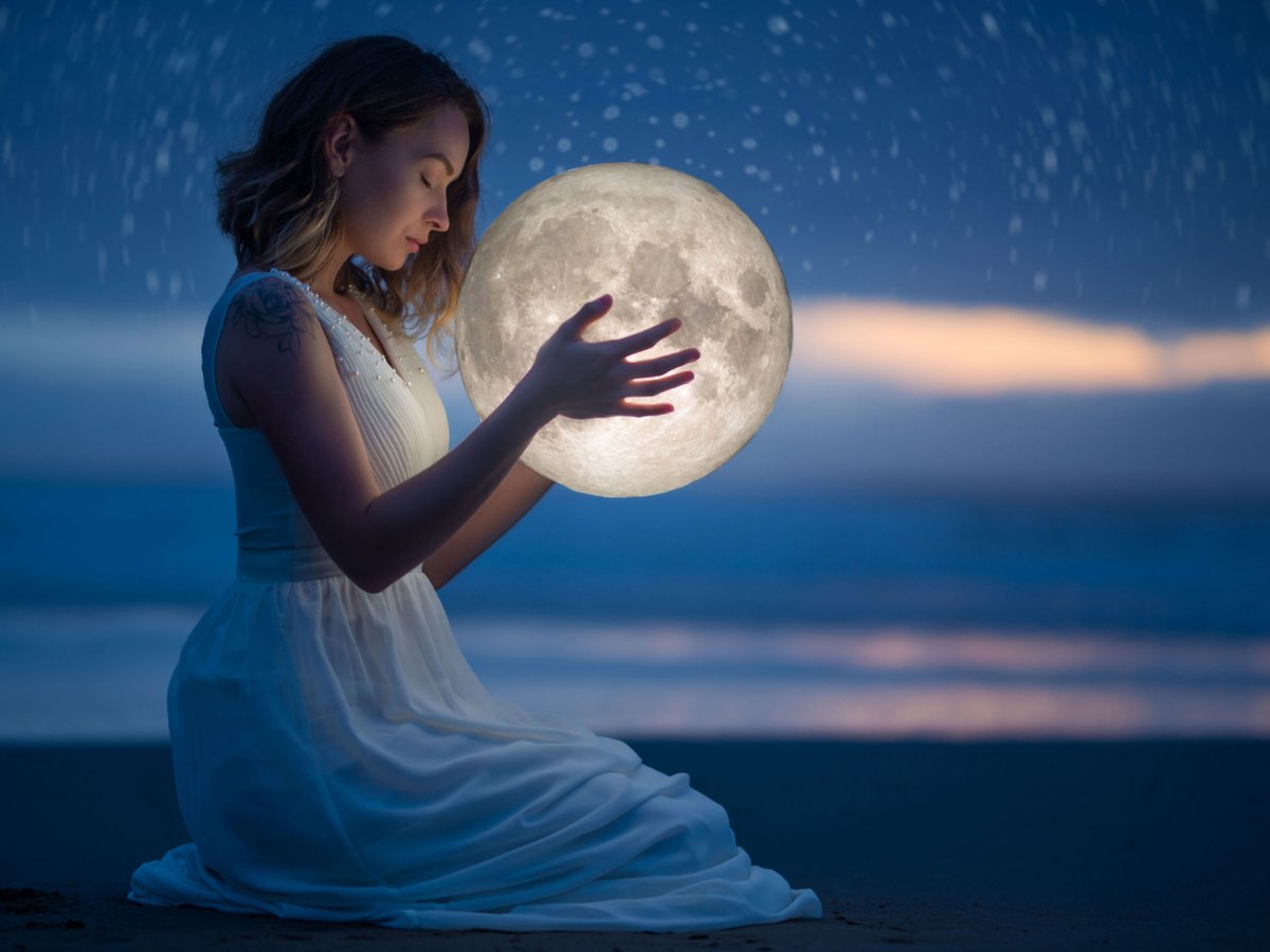 Frau im weißen Kleid bei Dämmerung am Strand hält Mond in der Hand
