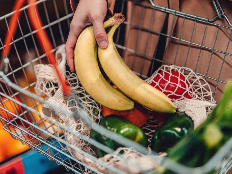 Frau legt Bananen in einen Einkaufskorb, in dem bereits viel Gemüse und andere Lebensmittel liegen.