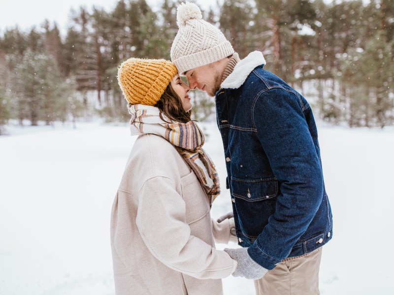 Ein junges Paar in Winterkleidung steht mit Stirn an Stirn lächelnd in einer schneebedeckten Waldlandschaft.