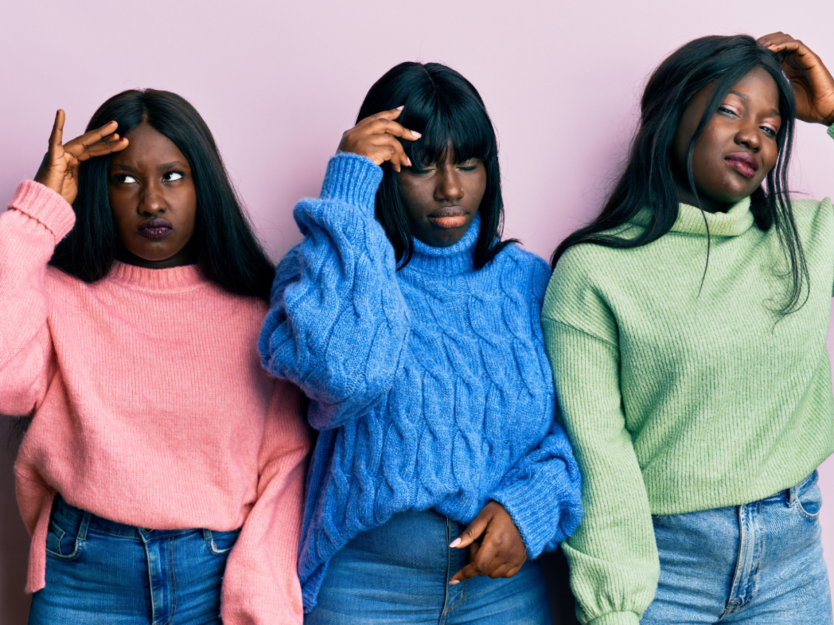 Drei junge Frauen mit langen schwarzen Haaren und ernsten Gesichtsausdrücken tragen bunte Strickpullover vor einer rosa Wand.