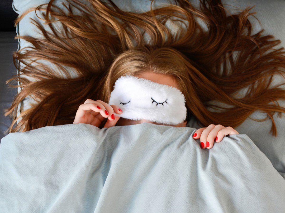 Junge Frau mit langen Haaren und roter Manik&uuml;re schl&auml;ft mit Schlafmaske im Bett und schaut unter der Schlafmaske hervor