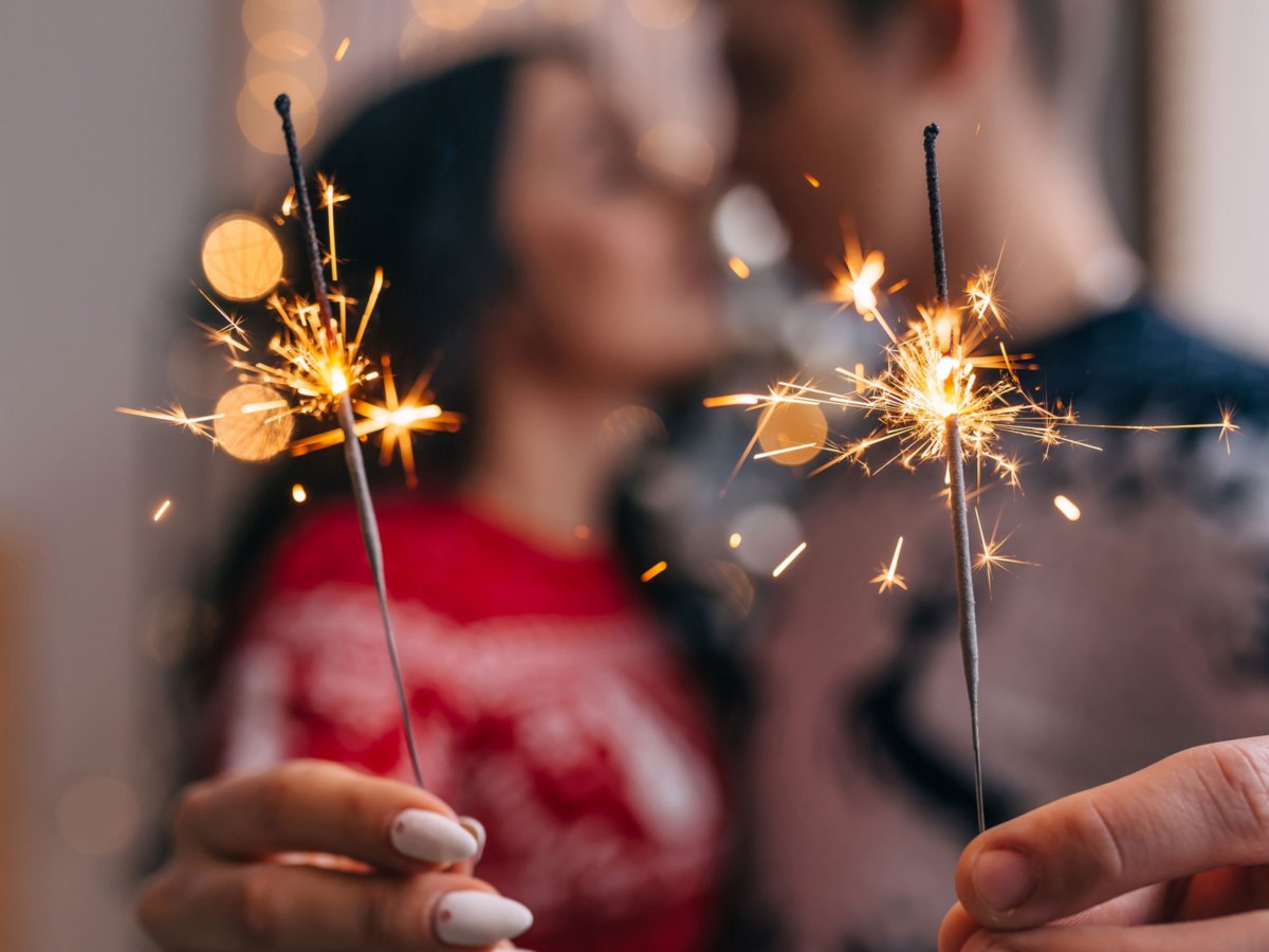 Frau und Mann in Wiehnachtspullovern stehen sich Nase an Nase und halten Wunderkerzen in die Kamera
