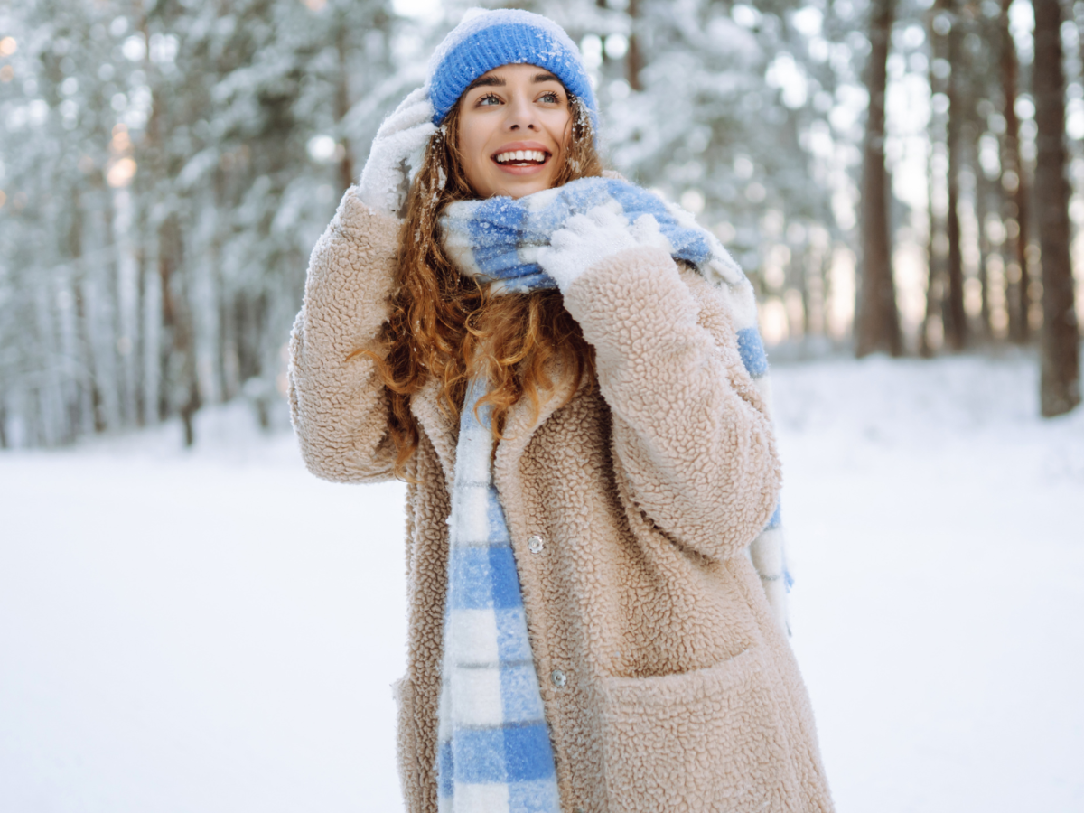 Junge Frau mit braunen Locken und blauer M&uuml;tze lacht fr&ouml;hlich im verschneiten Winterwald, eingeh&uuml;llt in einen beigen Mantel und karierten Schal.
