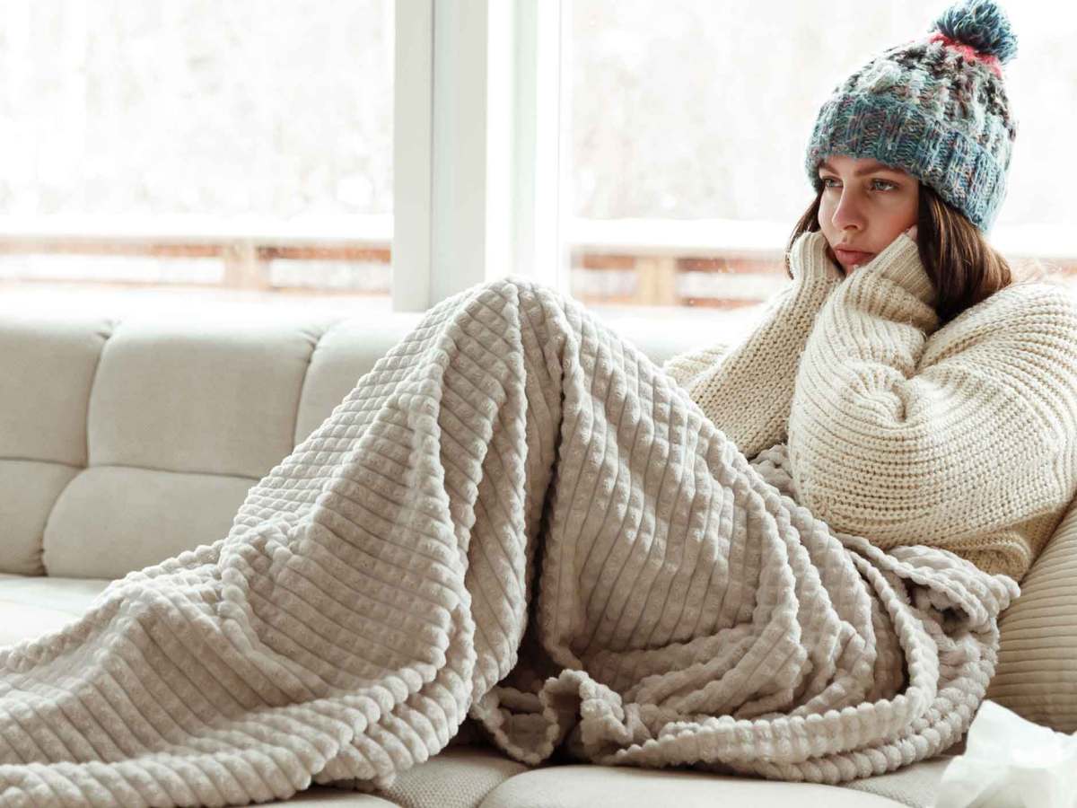 Frau sitzt eingepackt mit Decke und M&uuml;tze auf der Couch und schmollt.