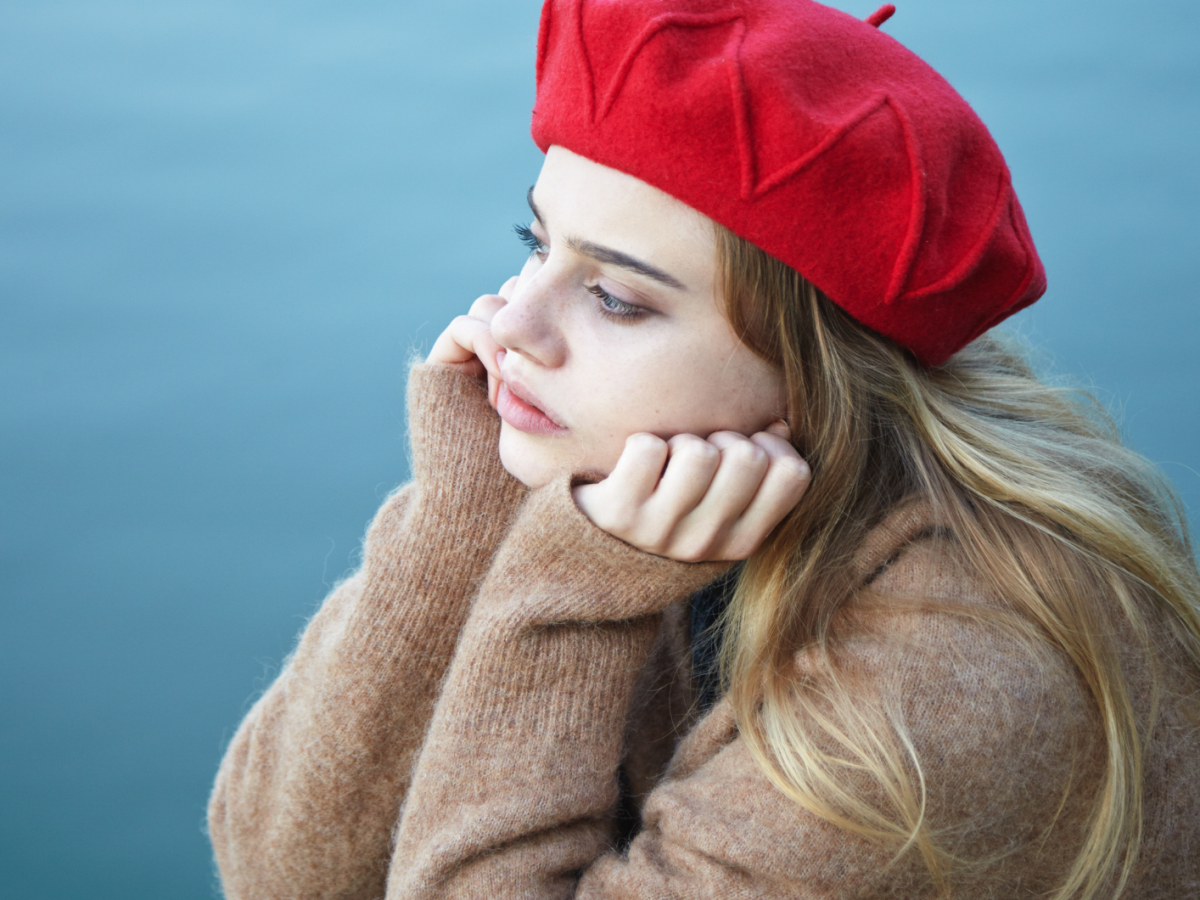 Junge blonde Frau mit rotem Barett und nachdenklichem Blick, gest&uuml;tzt auf ihre H&auml;nde, sitzt vor einem ruhigen, blauen Gew&auml;sser.