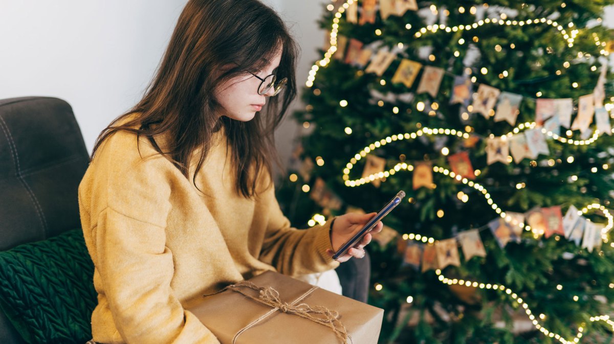 Teenagermädchen sitzt mit Geschenk auf dem Schoß vorm Weihnachtsbaum und schaut in ihr Handy.