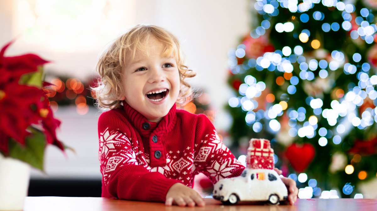 Junge, ca. 6 Jahre, lacht sehr glücklich, hinter ihm steht ein leuchtender Weihnachtsbaum.