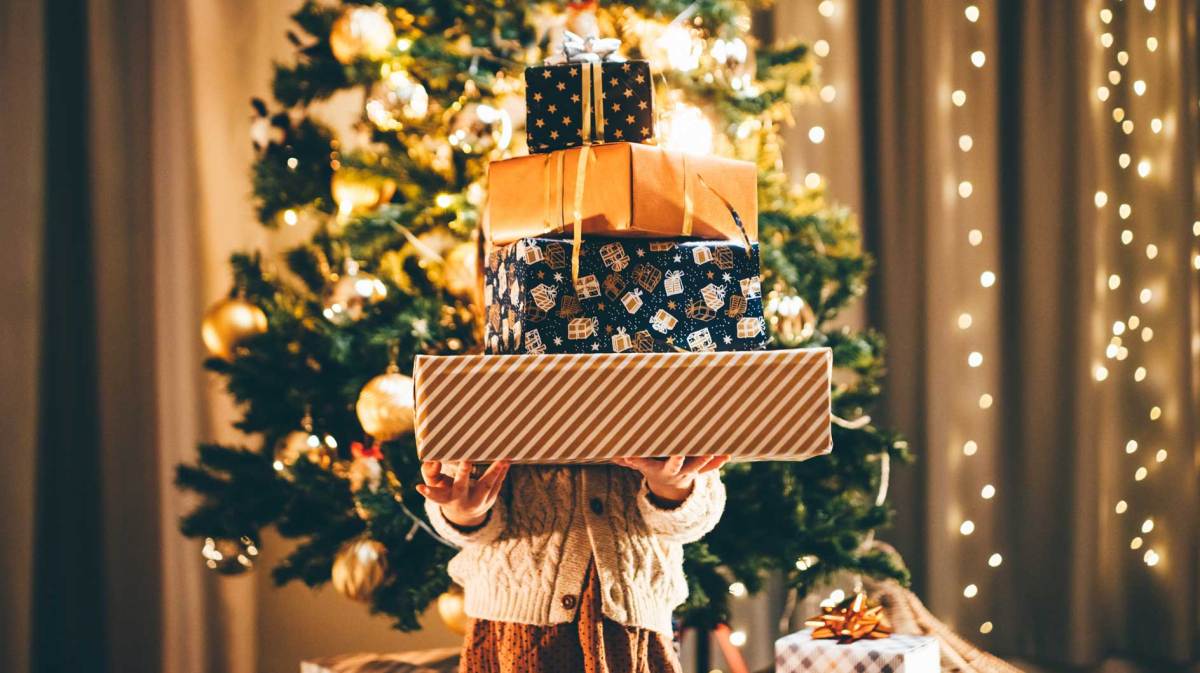 Kind steht vor dem beleuchteten Weihnachtsbaum und hält einen Berg Weihnachtsgeschenke und ist selbst nicht mehr zu sehen.