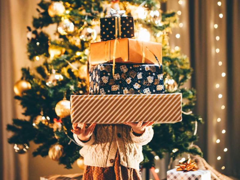 Kind steht vor dem beleuchteten Weihnachtsbaum und hält einen Berg Weihnachtsgeschenke und ist selbst nicht mehr zu sehen.
