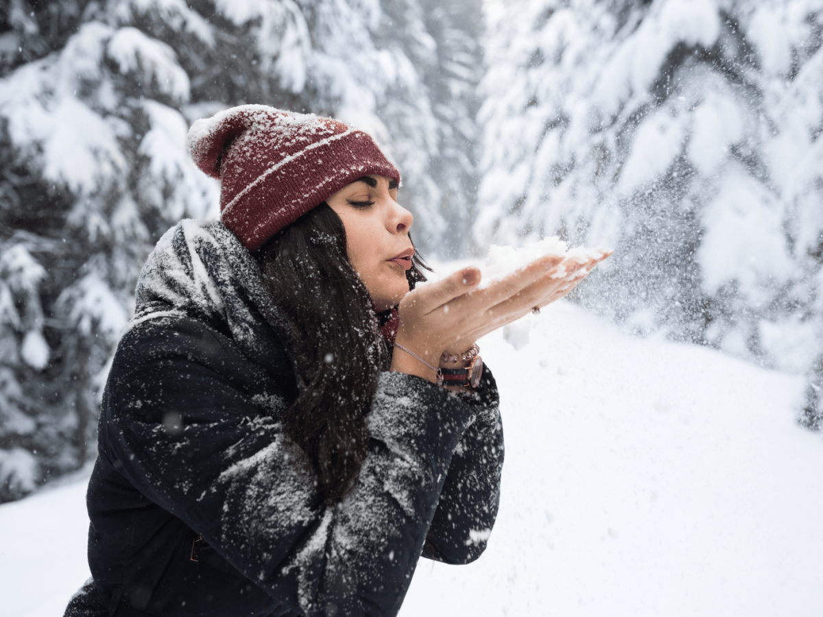 Junge Frau mit schwarzem Haar und roter M&uuml;tze pustet Schnee aus ihren H&auml;nden in einem verschneiten Wald voller Tannen.
