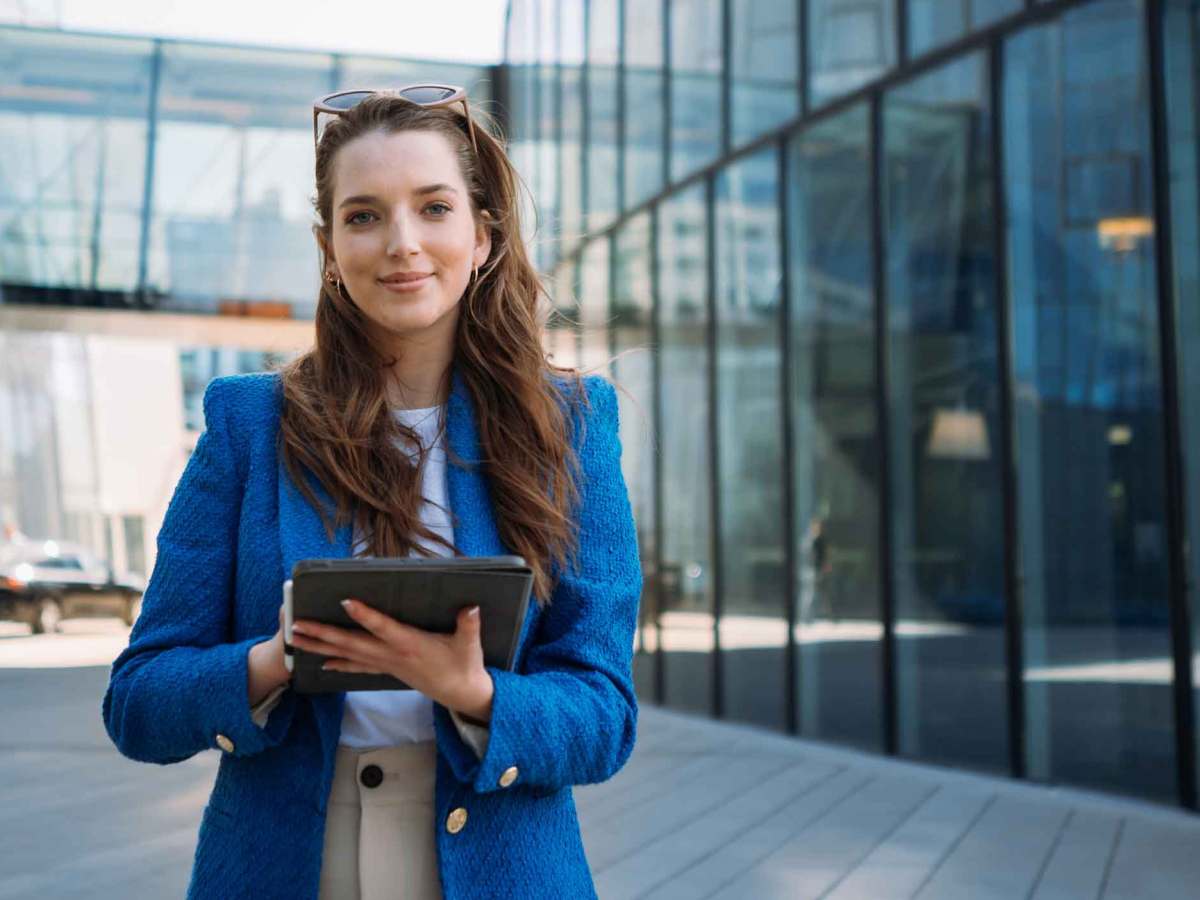 Frau mit braunen Haaren und einem blauen Blazer steht vor einem B&uuml;rogeb&auml;ude und h&auml;lt ein Tablet in den H&auml;nden.