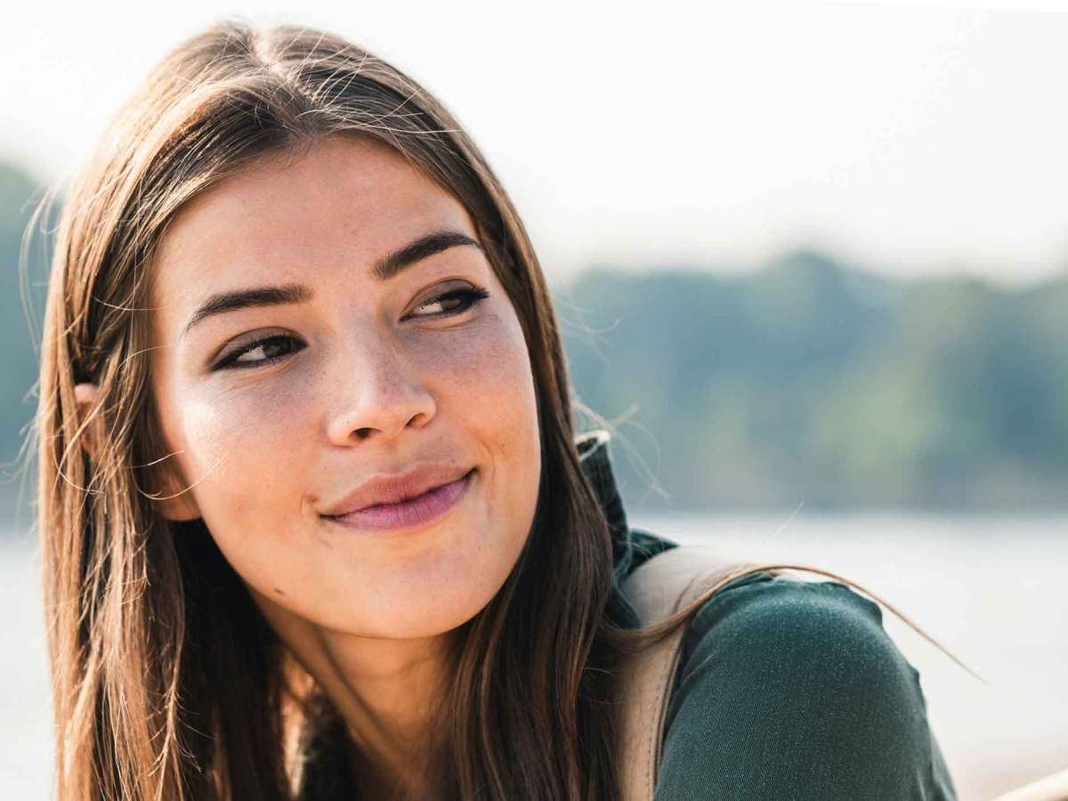 Junge Frau mit glatte dunklen Haaren sitzt drau&szlig;en am Wasser und blickt freundlich zur Seite.