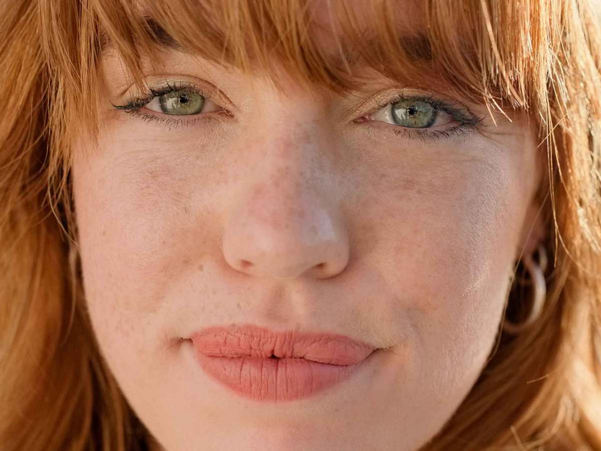 Nahaufnahme einer jungen Frau mit rotblonden Haaren und hellen Augen, die zurückhaltend lächelt.