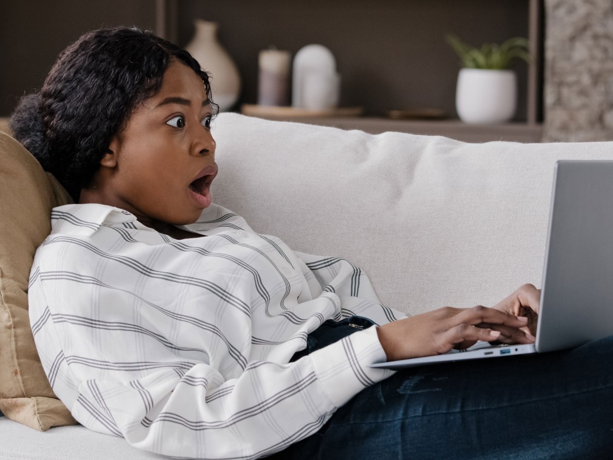 Junge Frau, die zu Hause auf der Couch liegt und in den Laptop schaut, um unerwartete schlechte Nachrichten zu lesen, &ouml;ffnet den Mund weit in &Uuml;berraschung und Schock und schaut auf Computer