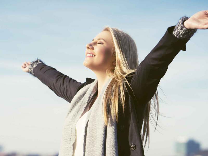 Blonde Frau mit Schal steht im Januar draußen und breitet zufrieden die Arme nach oben aus.