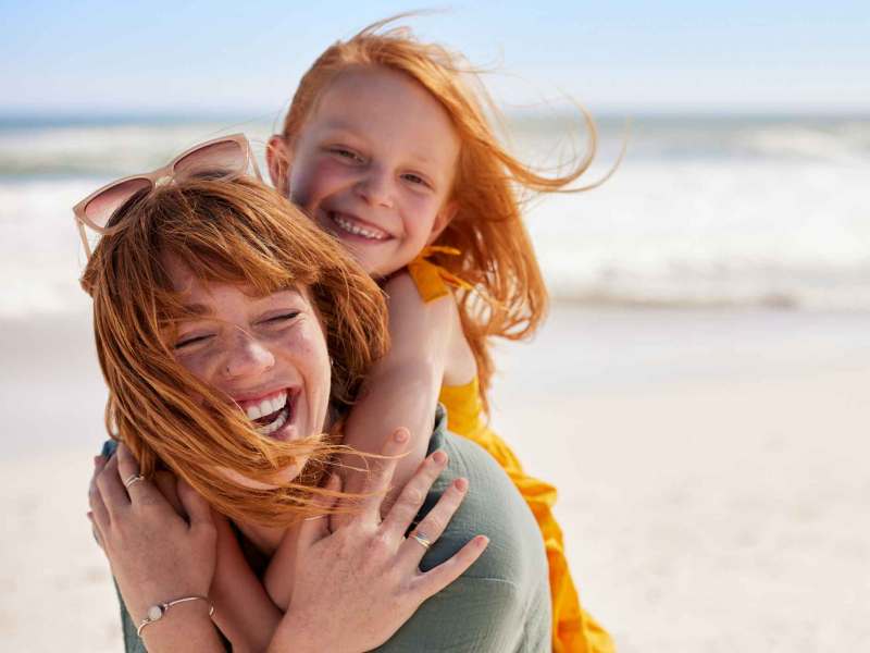 Eine lachende rothaarige Frau trägt ein rothaariges Mädchen in gelbem Kleid am sonnigen Strand, beide wirken ausgelassen.