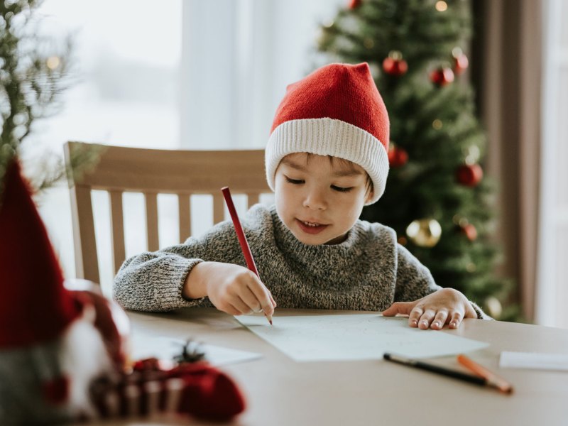 Kind sitzt am Tisch, trägt eine Weihnachtsmannmütze und malt einen Wunschzettel.