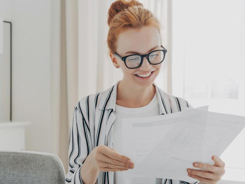 Frau mit Brille und einer strengen Frisur sitzt mit Dokumenten am Schreibtisch.