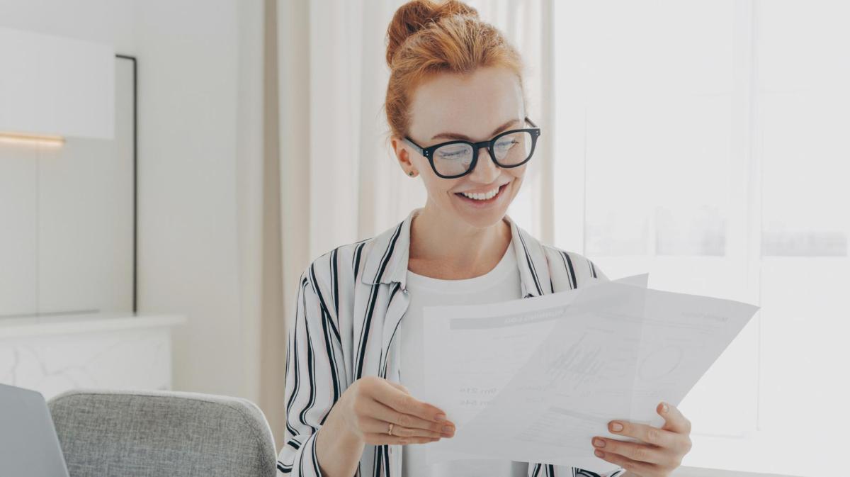 Frau mit Brille und einer strengen Frisur sitzt mit Dokumenten am Schreibtisch.