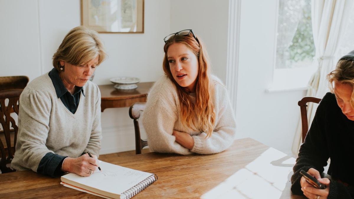Drei Personen sitzen an einem Holztisch in einem hellen Raum, eine ältere Frau zeichnet, während eine junge Frau mit roten Haaren spricht.