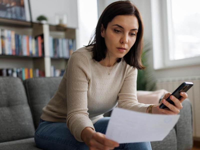 Eine junge Frau mit schulterlangem braunem Haar sitzt nachdenklich auf einem grauen Sofa, hält ein Dokument und schaut auf ihr Smartphone.