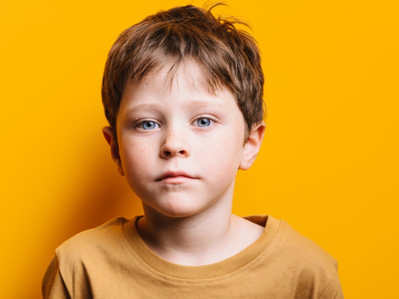Portr&auml;t eines Jungen, ca. 7 Jahre alt, der in einem gelben Shirt vor einer gelben Wand steht und ernst schaut.