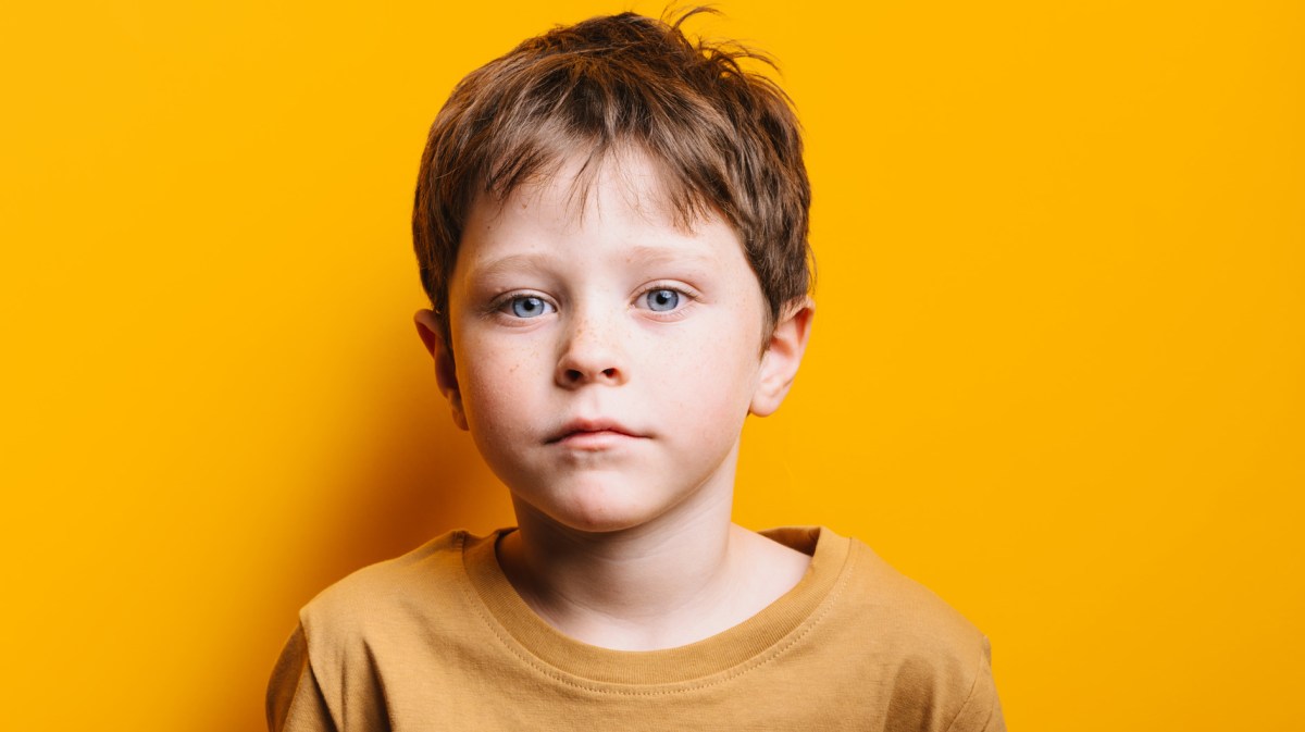Porträt eines Jungen, ca. 7 Jahre alt, der in einem gelben Shirt vor einer gelben Wand steht und ernst schaut.