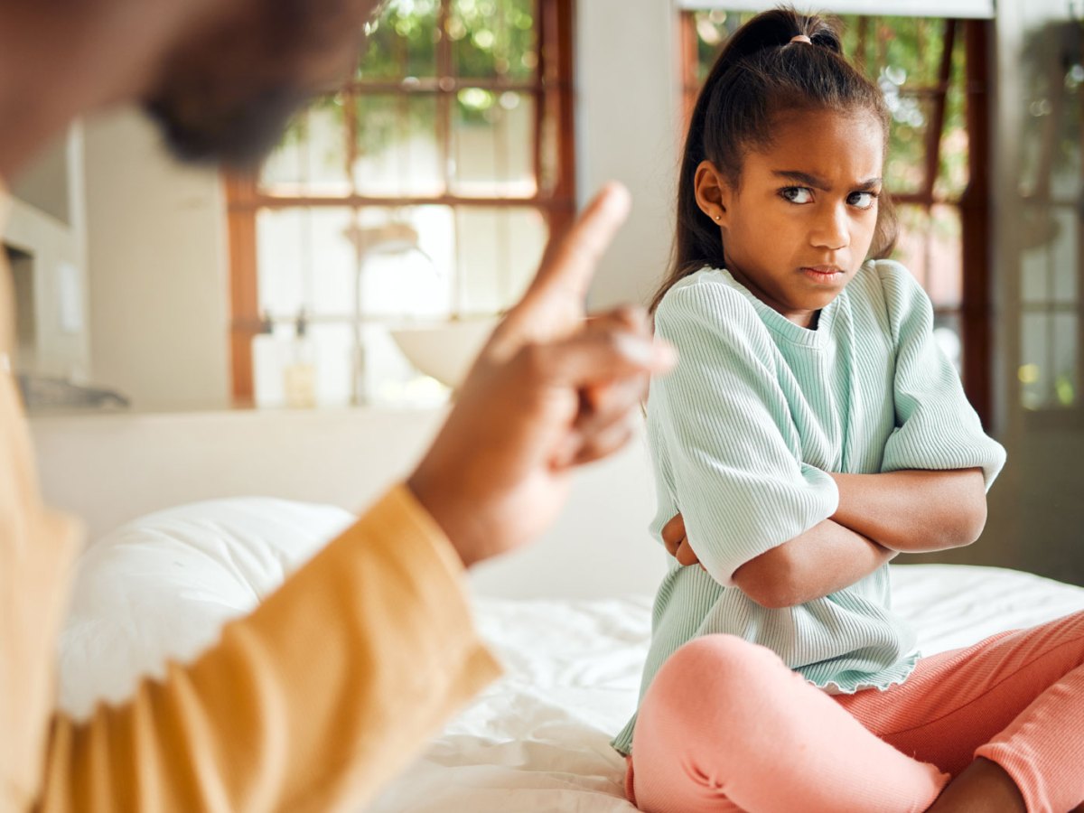 W&uuml;tendes M&auml;dchen sitzt auf ihrem Bett, verschr&auml;nkt die Arme und schaut ihren Vater b&ouml;se an.