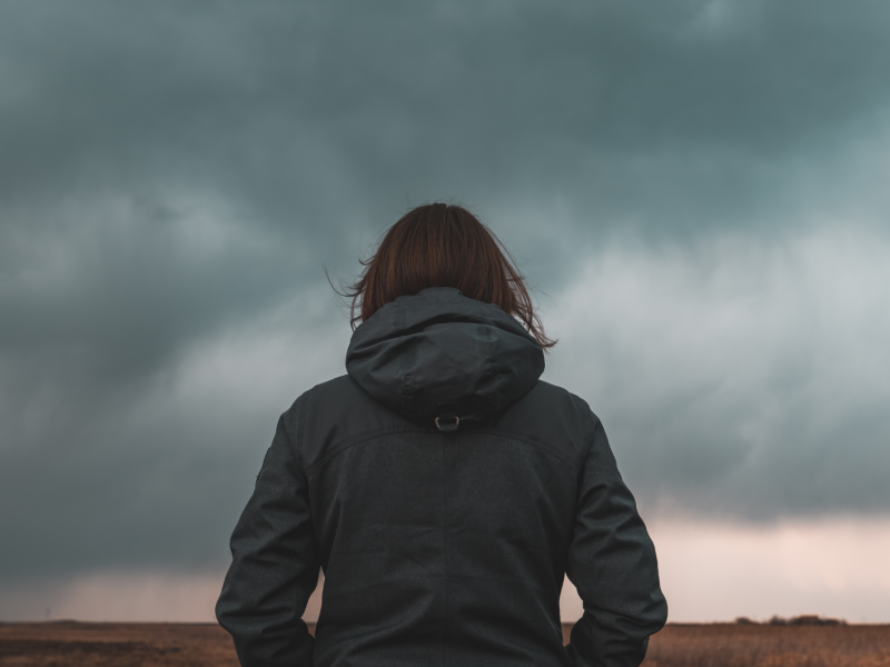 Frau steht mit dem Rücken zur Kamera vor einem düsteren Wolkenhimmel