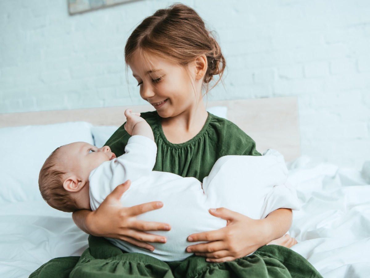 Gl&uuml;ckliches M&auml;dchen h&auml;lt ihre kleine Baby-Schwester im Arm, w&auml;hrend sie auf einem wei&szlig;en Bett sitzt.