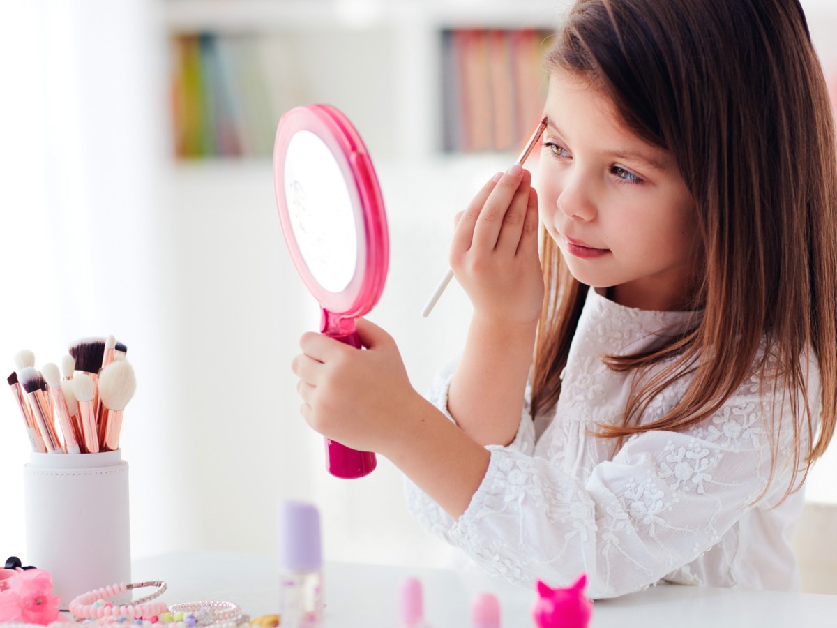 Kleines M&auml;dchen, ca. 6 bis 8 Jahre, sitzt mit einem Handspiegel am Tisch und tr&auml;gt mit einem Make-up-Pinsel Augen-Make-up auf.