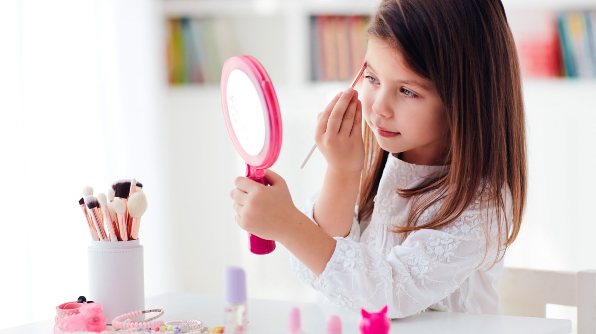 Kleines Mädchen, ca. 6 bis 8 Jahre, sitzt mit einem Handspiegel am Tisch und trägt mit einem Make-up-Pinsel Augen-Make-up auf.