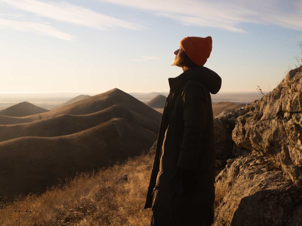 Eine Person mit orangefarbener M&uuml;tze und langem Mantel steht bei Sonnenuntergang auf einem Felsvorsprung und blickt &uuml;ber h&uuml;gelige Landschaft.