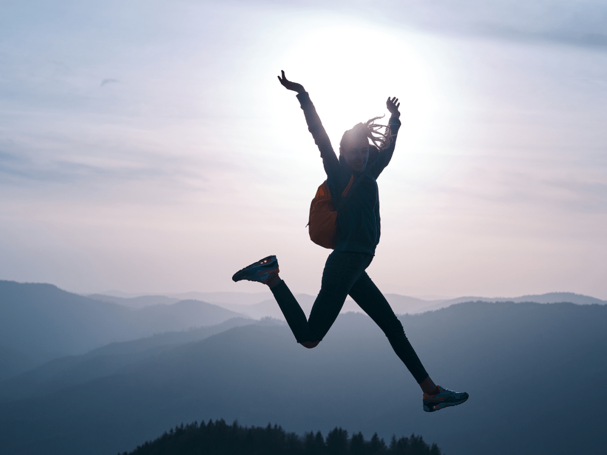 Frau mit Rucksack auf einem Berg, springt freudestrahlend in die Luft