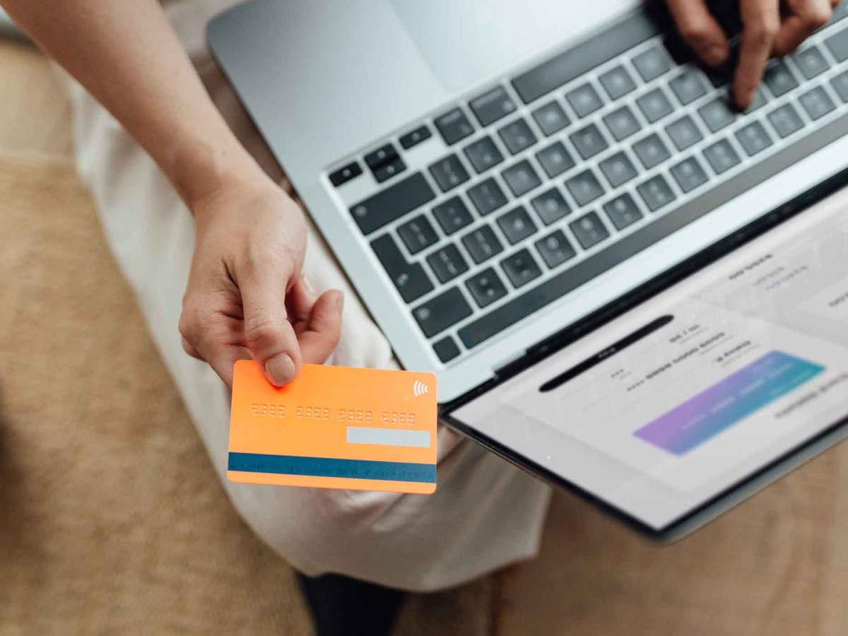 Eine Person in heller Kleidung h&auml;lt eine orange Kreditkarte vor einem Laptop mit ge&ouml;ffneter Zahlungsseite auf einem Teppich.