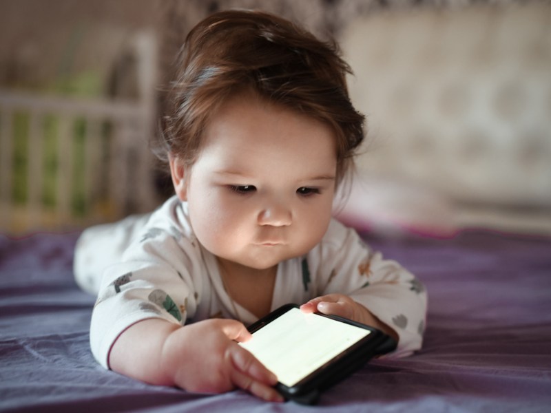 Baby liegt auf dem Bauch auf einem Bett und schaut auf einen leuchtenden Handybildschirm.